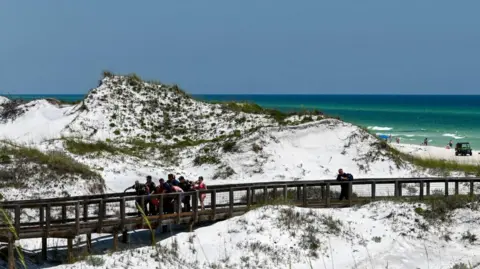 South Walton Fire District A beach with a walkway, where a crowd of rescuers are surrounding a person being carried on a stretcher